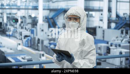 Female Worker Wearing Full-Body Protective Coveralls, Mask and Goggles at a Manufacturing Plant. Portrait of a Young Woman Specialists Using a Tablet Computer, Looking at Camera, Smiling Stock Photo