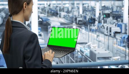 Young Female Engineer Using Laptop Computer with Green Screen Mock Up Display at an Electronics Factory. Automated Manufacturing Machines and AI Robots Are Monitored Online Stock Photo
