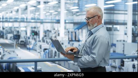 Electronics Manufacturing Technician Working on a Laptop Computer at a Factory Space. Specialist Developing, Maintaining, Fine-Tuning Programs for Machine Production and Assembly Stock Photo