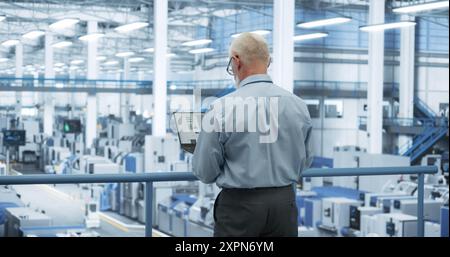 Middle Aged Engineer Monitoring and Analyzing Conditions at an Automated Modern Electronics Factory with Mobility Robots Working with the Help of Artificial Intelligence Software Stock Photo