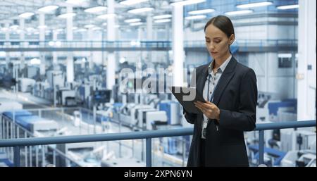 Thoughtful Female Engineer Monitoring and Analyzing Conditions at a Modern Tech Electronics Factory with Automated Robot Arms Working with the Help of Tablet Computer Stock Photo