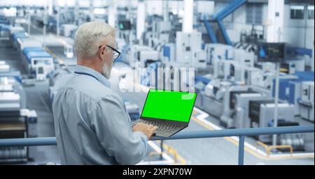 Technician Using Laptop Computer with Green Screen Chromakey Mock Up Display and Looking Around a Factory Facility with Modern Machines Producing Electronic Components for Tech Industries Stock Photo