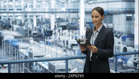 Portrait of a Beautiful Happy Engineer Using Laptop Computer and Looking Around a Factory Facility with Automated Robotic Arms Producing Modern Electronic Components for Different Industries Stock Photo