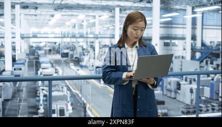 Thoughtful Asian Engineer Monitoring and Analyzing Work at a Modern Electronics Factory with Automated Robot Hands Making Devices. Female Work Colleague Passing By with a Tablet Device Stock Photo