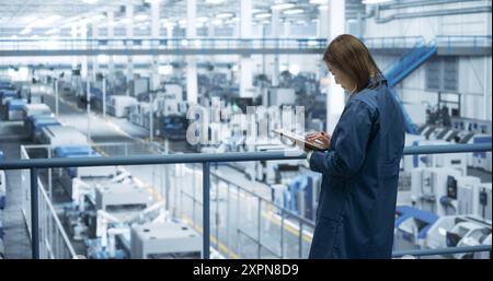 Experienced Female Engineer Standing on a Platform, Using Tablet Computer and Overlooking Production at a Modern Automated Electronics Manufacture with AI Support. Factory Manager at Work Stock Photo