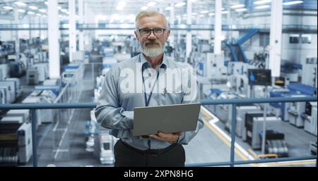 Portrait of a Handsome Middle Aged Engineer Wearing Glasses, Using Laptop Computer and Looking at Camera. Technician in High-VIsibility Vest Working in a Modern Technology Factory Stock Photo