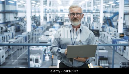 Portrait of a Senior Technician with a Beard and Glasses Using Laptop Computer and Looking at Camera. Industrial Male Specialist Wearing a Protective Vest, Working in a Modern Electronics Factory Stock Photo