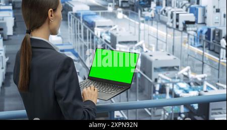 Female Technician Using Laptop Computer with Green Screen Chroma Key Mock Up Display and Looking Around a Factory Facility with Robotic Arms Producing Modern Electronic Components for Tech Industries Stock Photo