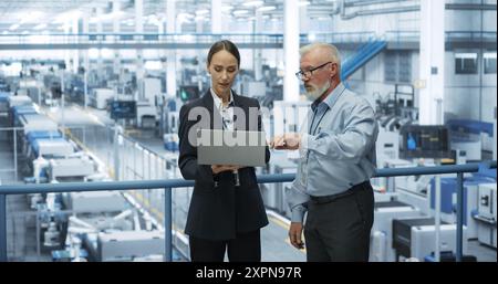 Experienced Male and Female Engineers Standing on a Platform at a Modern Industrial Factory, Using Laptop Computer and Discussing Production of Modern Electronics for Artificial Intelligence Machines Stock Photo