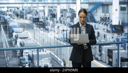 Portrait of a Caucasian Female Engineer Using Laptop Computer and Looking Around a Factory Facility with Robotic Arms Producing Modern Electronic Components for Tech Industries Stock Photo
