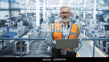 Portrait of a Senior Technician with a Beard and Glasses Using Laptop Computer and Looking at Camera. Industrial Male Specialist Wearing a Protective Vest, Working in a Modern Electronics Factory Stock Photo
