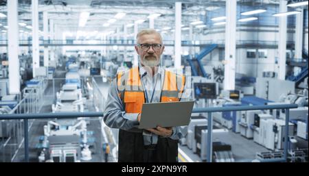 Portrait of a Handsome Middle Aged Engineer Wearing Glasses, Using Laptop Computer and Looking at Camera. Technician in High-VIsibility Vest Working in a Modern Technology Factory Stock Photo