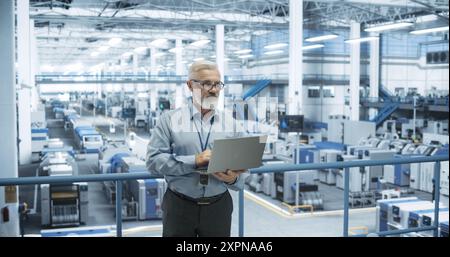 Senior Engineer Standing on a Platform, Using Laptop Computer at an Electronics Factory. Machines are Undergoing Maintenance, Specialist Monitoring the Progress Through Online Software Stock Photo