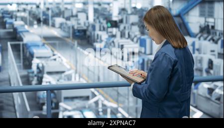 Experienced Female Engineer Standing on a Platform, Using Tablet Computer and Overlooking Production at a Modern Automated Electronics Manufacture with AI Support. Factory Manager at Work Stock Photo