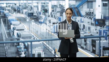 Electronics Manufacturing Technician Working on a Laptop Computer at a Factory Space. Female Specialist Developing, Maintaining, Upgrading Programs for Machine Production and Assembly Stock Photo
