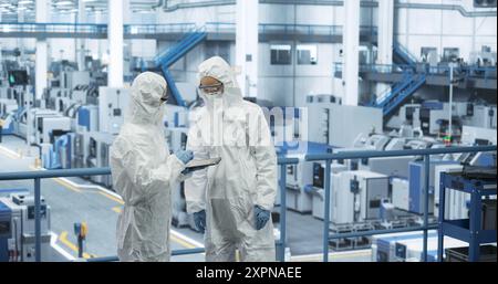 Two Female Technicians in Protective Coveralls Having a Conversation and Using Tablet Computer. Specialists Monitoring, Analyzing Conditions at a Modern Electronics Factory with Automated Robots Stock Photo