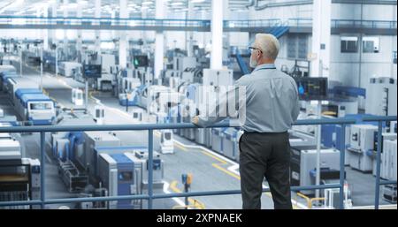 Industrial Engineer Working at a Technology Factory. Middle Aged Manager Standing on a Platform at Work, Looking at the Production Manufacturing Complex. Automated Robots Moving Around the Hall Stock Photo