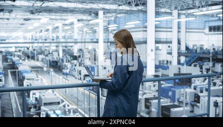 Experienced Female Engineer Standing on a Platform, Using Laptop Computer and Overlooking Production at a Modern Automated Electronics Manufacture with AI Support. Factory Manager at Work Stock Photo