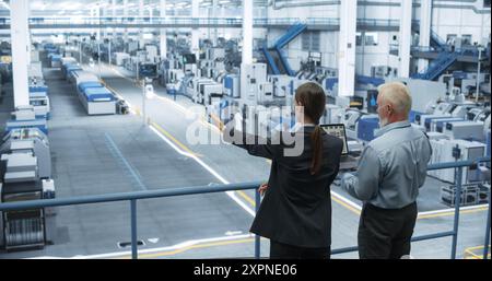 Male Engineer and Female Project Manager Standing on a Platform, Talking, Using Laptop Computer and Monitoring Production at a Modern Automated Electronics Manufacture with AI Robotic Arms Stock Photo