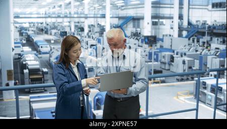 Factory Managers Working on Laptop at a Modern Manufacture with Autonomous Robot Hands. Female and Male Specialists Monitoring Technical Data on a Computer, Analyzing Reports and Planning Updates Stock Photo
