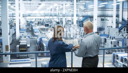 Portrait of a Middle Aged Engineer and Happy Asian Scientist Using Laptop Computer and Talking in a Factory Facility with Equipment Producing Modern Electronic Components for Different Industries Stock Photo