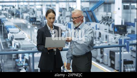 Experienced Male and Female Engineers Standing on a Platform at a Modern Industrial Factory, Using Laptop Computer and Discussing Production of Modern Electronics for Artificial Intelligence Machines Stock Photo