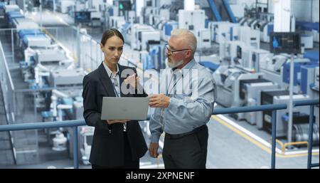 Experienced Male and Female Engineers Standing on a Platform at a Modern Industrial Factory, Using Laptop Computer and Discussing Production of Modern Electronics for Artificial Intelligence Machines Stock Photo