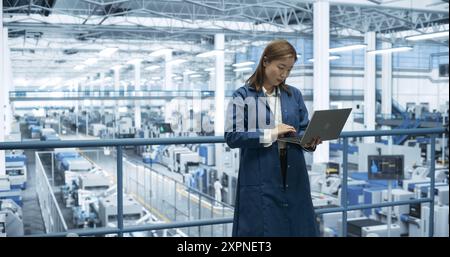 Electronics Manufacturing Technician Working on a Laptop Computer at a Factory Space. Asian Female Specialist Developing, Maintaining, Fine-Tuning Programs for Machine Production and Assembly Stock Photo