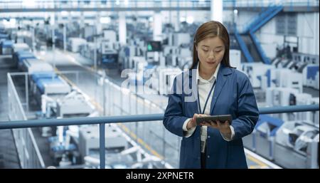 Japanese Factory Manager Working on a Tablet at a Futuristic Industrial Manufacture with Autonomous AI Robots. Specialist Monitoring All the Data on a Device, Checking Reports and Planning Research Stock Photo