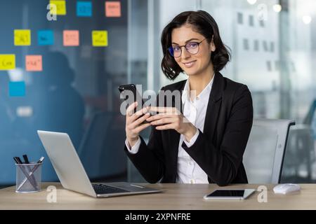Confident businesswoman in glasses using phone in modern office. Smiling professional working on laptop surrounded by colorful sticky notes, showcasing productivity and modern business environment. Stock Photo