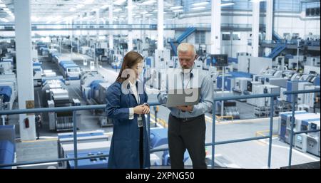 Factory Managers Working on Laptop at a Modern Manufacture with Autonomous Robot Hands. Female and Male Specialists Monitoring Technical Data on a Computer, Analyzing Reports and Planning Updates Stock Photo