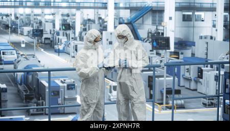 Two Female Technicians in Protective Coveralls Having a Conversation and Using Tablet Computer. Specialists Monitoring, Analyzing Conditions at a Modern Electronics Factory with Automated Robots Stock Photo
