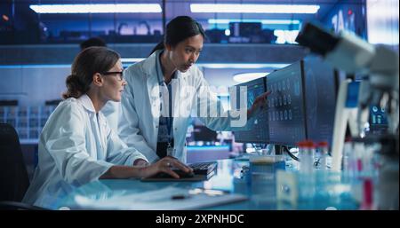 Asian And Caucasian Female Doctors Talking In Medical Research Center And Using Desktop Computer To Analyze MRI Scans Of Brain. Surgeon And Neuroscientist Discussing Solutions For Treating Cancer. Stock Photo