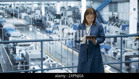 Japanese Factory Manager Working on a Tablet at a Futuristic Industrial Manufacture with Autonomous AI Robots. Specialist Monitoring All the Data on a Device, Checking Reports and Planning Research Stock Photo