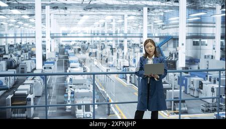 Shot in an Electronics Factory. Japanese Project Manager Working on Laptop at a Futuristic Industrial Manufacture with AI Robotic Arms. Specialist Monitoring Production on a Computer Stock Photo