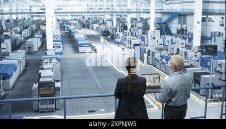 Male Engineer and Female Project Manager Standing on a Platform, Talking, Using Laptop Computer and Monitoring Production at a Modern Automated Electronics Manufacture with AI Robotic Arms Stock Photo