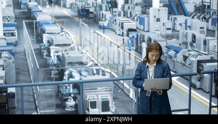 Japanese Factory Specialist Working on Laptop at a Futuristic Industrial Manufacture with Autonomous AI Robots. Project Manager Monitoring Data on a Computer, Checking Reports and Planning Research Stock Photo