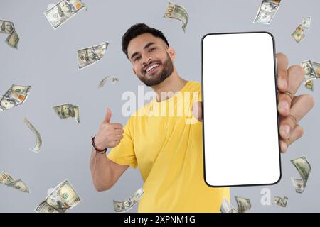 Happy man showing mobile phone with empty screen under money rain on grey background Stock Photo