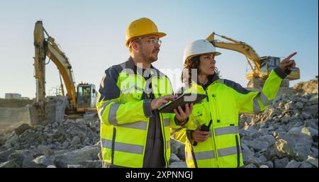 Caucasian Male Civil Engineer Talking To Hispanic Female Urban Planner And Using Tablet On Construction Site of New Apartment Building. Real Estate Developers Discussing Project, Excavators Working. Stock Photo