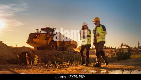 Caucasian Male Architect And Hispanic Female Urban Planner Walking On Construction Site With Laptop Computer And Talking About New Real Estate Project. Construction Truck Driving By. Golden Hour Stock Photo