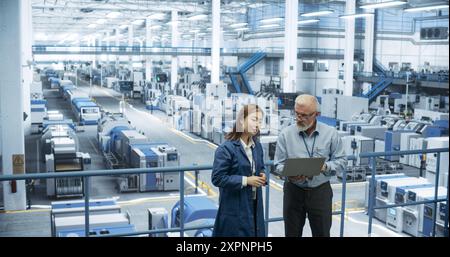 Portrait of a Middle Aged Engineer and Positive Asian Scientist Using Laptop Computer and Talking in a Factory Facility with Equipment Producing Modern Electronic Components for Different Industries Stock Photo