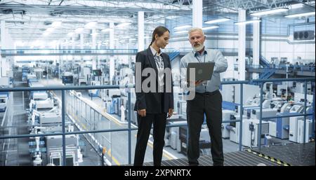 Portrait of a Middle Aged Engineer and Female Scientist Using Laptop Computer and Talking in a Factory Facility with Equipment Producing Modern Technical Devices for Different Industries Stock Photo