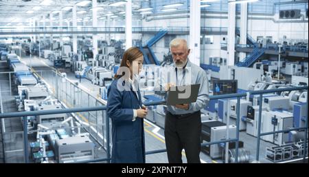 Factory Managers Working on Laptop at a Modern Manufacture with Autonomous Robot Hands. Female and Male Specialists Monitoring Technical Data on a Computer, Analyzing Reports and Planning Updates Stock Photo