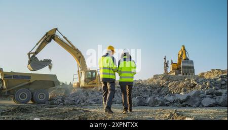 Back View Of Hispanic Female Project Manager And Caucasian Male Civil Engineer Walking and Discussing Real Estate Project On Construction Site. Excavators Digging The Ground For Building Foundation Stock Photo
