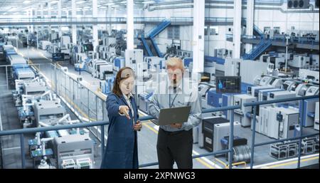 Portrait of a Middle Aged Engineer and Happy Asian Scientist Using Laptop Computer and Talking in a Factory Facility with Equipment Producing Modern Electronic Components for Different Industries Stock Photo