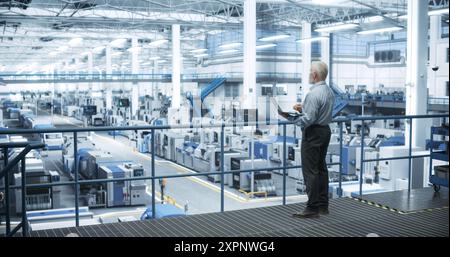 Senior Engineer Standing on a Platform, Using Laptop Computer at an Electronics Factory. Machines are Undergoing Maintenance, Specialist Monitoring the Progress Through Online Software Stock Photo