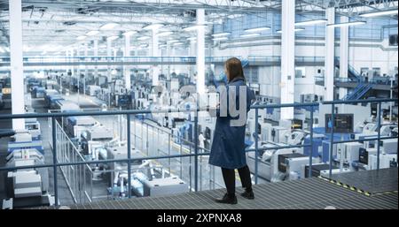 Japanese Factory Manager Working on Laptop at a Futuristic Industrial Manufacture with Autonomous AI Robots. Specialist Monitoring All the Data on a Computer, Checking Reports and Planning Research Stock Photo