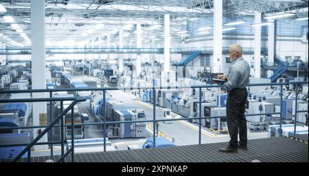 Experienced Male Engineer Standing on a Platform, Using Laptop Computer and Overlooking Production at a Modern Automated Electronics Manufacture with AI Support. Factory Executive at Work Stock Photo