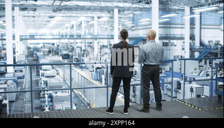 Male Engineer and Female Project Manager Standing on a Platform, Talking, Using Laptop Computer and Monitoring Production at a Modern Automated Electronics Manufacture with AI Robotic Arms Stock Photo