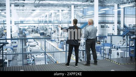 Male Engineer and Female Project Manager Standing on a Platform, Talking, Using Laptop Computer and Monitoring Production at a Modern Automated Electronics Manufacture with AI Robotic Arms Stock Photo